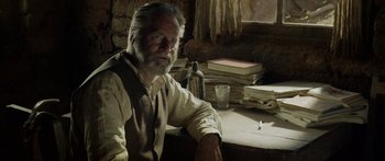 Movie still from “Blackthorn” (2011), directed by Mateo Gil – An older man sitting at a table with papers and a bottle; Medium shot, Over the shoulder angle