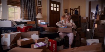 Movie still from “Senior Year” (2022), directed by Alex Hardcastle – A woman sitting on a chair in a room filled with lots of boxes; Wide shot, High angle