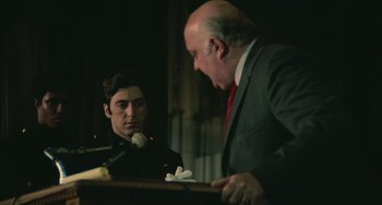 Movie still from “Serpico” (1973), directed by Sidney Lumet – A man in a suit and tie talking to another man in front of a microphone; Medium shot, Over the shoulder angle