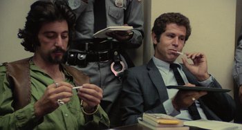 Movie still from “Serpico” (1973), directed by Sidney Lumet – Two men sitting at a table and one of the men is smoking a cigarette and the other is holding a cell phone; Medium shot, Low angle