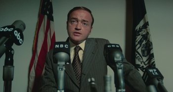 Movie still from “Serpico” (1973), directed by Sidney Lumet – A man standing in front of microphones in front of an american flag; Close Up shot, Low angle