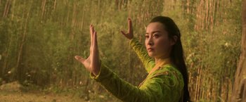 Movie still from “Shang-Chi and the Legend of the Ten Rings” (2021), directed by Destin Daniel Cretton – A woman in a yellow and green jacket holding her hands up in front of a forest; Medium shot, Low angle