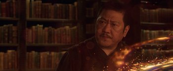Movie still from “Shang-Chi and the Legend of the Ten Rings” (2021), directed by Destin Daniel Cretton – A man in a dark room with a book shelf in the background; Close Up shot, Over the shoulder angle