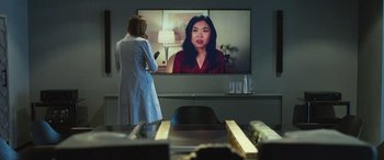 Movie still from “Shazam!” (2019), directed by David F. Sandberg – A woman in a white lab coat standing in front of a television; Medium shot, Over the shoulder angle