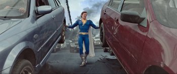 Movie still from “Shazam! Fury of the Gods” (2023), directed by David F. Sandberg – A man dressed as captain marvel standing next to two cars; Wide shot, Low angle