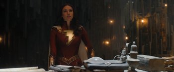 Movie still from “Shazam! Fury of the Gods” (2023), directed by David F. Sandberg – A woman dressed as captain marvel standing in front of a table; Medium shot, Over the shoulder angle