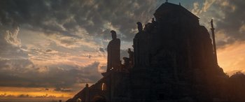 Movie still from “Shazam! Fury of the Gods” (2023), directed by David F. Sandberg – The sun is setting behind a building with statues; Extreme Wide shot, Low angle
