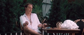 Movie still from “Shelter in Place” (2021), directed by Connor Martin – A man sitting at an outdoor table holding a cell phone; Medium shot, Low angle