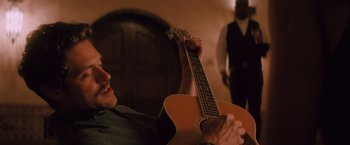 Movie still from “Shelter in Place” (2021), directed by Connor Martin – A man holding a guitar in a room with a man standing behind him; Medium shot, Over the shoulder angle