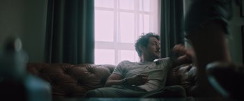 Movie still from “Shelter in Place” (2021), directed by Connor Martin – A man sitting on top of a couch in a living room; Medium shot, Low angle