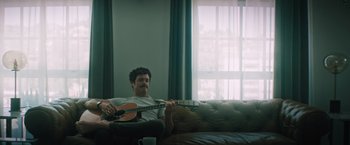 Movie still from “Shelter in Place” (2021), directed by Connor Martin – A man sitting on a couch holding a guitar; Medium shot, Low angle