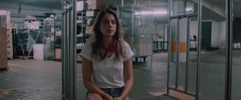 Movie still from “Shelter in Place” (2021), directed by Connor Martin – A woman sitting in a warehouse wearing shorts and a white shirt; Medium shot, Low angle