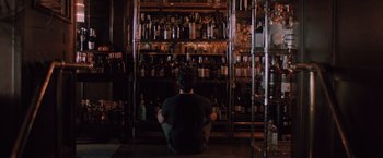 Movie still from “Shelter in Place” (2021), directed by Connor Martin – A man sitting in front of a shelf filled with bottles of alcohol; Medium shot, High angle