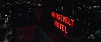 Movie still from “Shelter in Place” (2021), directed by Connor Martin – A neon sign is lit up on the side of a building; Extreme Wide shot, High angle