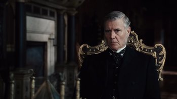 Movie still from “Sherlock Holmes” (2009), directed by Guy Ritchie – An older man wearing a suit and tie in front of an ornate chair; Close Up shot, Over the shoulder angle