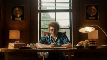 Movie still from “Bohemian Rhapsody” (2018), directed by Bryan Singer – An older woman sitting at a table writing on a piece of paper in front of a window; Medium shot, Low angle