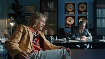 Movie still from “Bohemian Rhapsody” (2018), directed by Bryan Singer – A man sitting on a couch in front of a desk; Medium shot, Over the shoulder angle