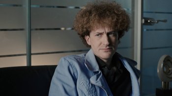 Movie still from “Bohemian Rhapsody” (2018), directed by Bryan Singer – A man sitting down wearing a blue jacket; Close Up shot, Over the shoulder angle