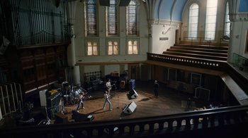 Movie still from “Bohemian Rhapsody” (2018), directed by Bryan Singer – A group of people standing in a large room with microphones; Extreme Wide shot, High angle