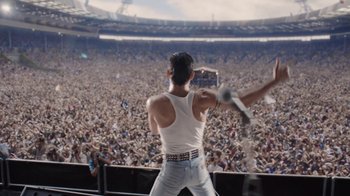 Movie still from “Bohemian Rhapsody” (2018), directed by Bryan Singer – A man in white shirt and white vest holding a microphone in front of a crowd; Extreme Wide shot, Low angle