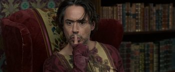 Movie still from “Sherlock Holmes: A Game of Shadows” (2011), directed by Guy Ritchie – A person sitting in a chair holding his finger to his mouth; Close Up shot, Over the shoulder angle