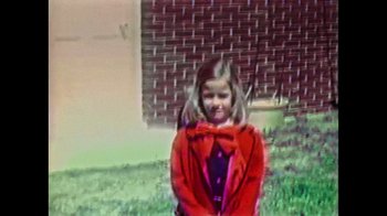 Movie still from “Sheryl” (2022), directed by Amy Scott – A little girl wearing a red jacket and a red bow tie; Medium shot, High angle