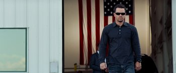 Movie still from “Shooter” (2007), directed by Antoine Fuqua – A man in a black shirt is walking in front of an american flag; Medium shot, Low angle