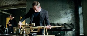 Movie still from “Shooter” (2007), directed by Antoine Fuqua – A man in a suit and tie is holding a wooden gun; Medium shot, High angle