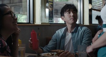 Movie still from “Shortcomings” (2023), directed by Randall Park – A man sitting in front of a plate of food; Medium shot, Over the shoulder angle