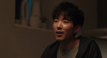 Movie still from “Shortcomings” (2023), directed by Randall Park – A young man is smiling while sitting in a room; Close Up shot, Over the shoulder angle