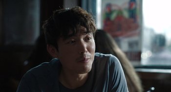 Movie still from “Shortcomings” (2023), directed by Randall Park – A young man with dark hair sitting in front of a window; Close Up shot, Over the shoulder angle