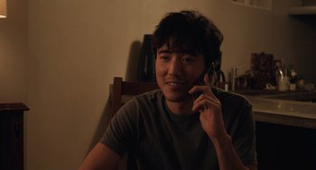 Movie still from “Shortcomings” (2023), directed by Randall Park – A man sitting in a chair talking on a cell phone; Close Up shot, Over the shoulder angle