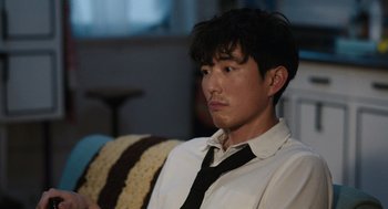 Movie still from “Shortcomings” (2023), directed by Randall Park – A man sitting on top of a couch wearing a tie; Close Up shot, Over the shoulder angle