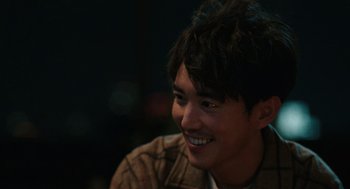 Movie still from “Shortcomings” (2023), directed by Randall Park – A young man smiles while sitting in a room; Close Up shot, Over the shoulder angle