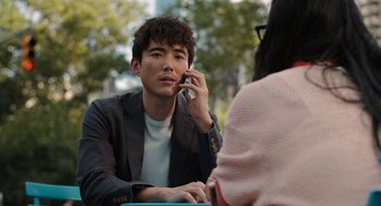 Movie still from “Shortcomings” (2023), directed by Randall Park – A man talking on a cell phone while sitting at a table; Medium shot, Over the shoulder angle