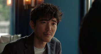 Movie still from “Shortcomings” (2023), directed by Randall Park – A man with a beard and a jacket is sitting in front of a blue wall; Close Up shot, Over the shoulder angle