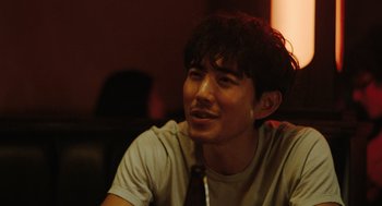 Movie still from “Shortcomings” (2023), directed by Randall Park – A man with a beard and a tie sitting in a room; Close Up shot, Over the shoulder angle