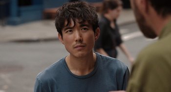 Movie still from “Shortcomings” (2023), directed by Randall Park – A man with a beard and a blue t - shirt is standing outside; Close Up shot, Over the shoulder angle