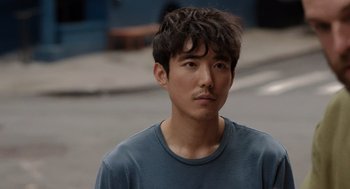 Movie still from “Shortcomings” (2023), directed by Randall Park – A man with a beard and a blue t - shirt is standing outside; Close Up shot, Over the shoulder angle