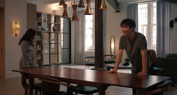 Movie still from “Shortcomings” (2023), directed by Randall Park – A man standing at a large wooden dining room table; Wide shot, Over the shoulder angle