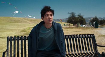 Movie still from “Shortcomings” (2023), directed by Randall Park – A man sitting on top of a wooden park bench; Medium shot, Over the shoulder angle