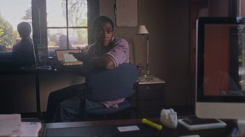 Movie still from “Showing Up” (2022), directed by Kelly Reichardt – A man sitting at a desk holding a remote control; Medium shot, Over the shoulder angle