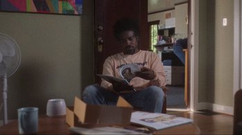 Movie still from “Showing Up” (2022), directed by Kelly Reichardt – A man sitting at a table reading a book; Medium shot, Over the shoulder angle