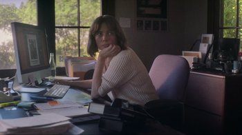 Movie still from “Showing Up” (2022), directed by Kelly Reichardt – A woman sitting at a table with a camera in front of a window; Medium shot, Over the shoulder angle