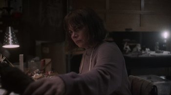 Movie still from “Showing Up” (2022), directed by Kelly Reichardt – A woman sitting at a table in a dark room; Medium shot, Low angle