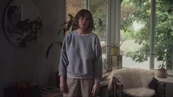 Movie still from “Showing Up” (2022), directed by Kelly Reichardt – A woman standing in a living room in front of a window; Medium shot, Over the shoulder angle