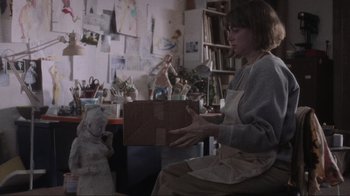 Movie still from “Showing Up” (2022), directed by Kelly Reichardt – A woman sitting in front of a statue in a room; Medium shot, Over the shoulder angle