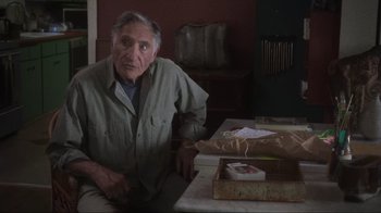Movie still from “Showing Up” (2022), directed by Kelly Reichardt – An older man sitting at a table in front of an open box; Medium shot, Over the shoulder angle
