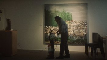 Movie still from “Showing Up” (2022), directed by Kelly Reichardt – A man is vacuuming in front of a painting; Wide shot, High angle