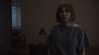 Movie still from “Showing Up” (2022), directed by Kelly Reichardt – A woman standing in a room with a lamp in the background; Medium shot, Over the shoulder angle